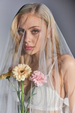 Beyaz peçeli, sarı saçlı, elinde çiçeklerle kameraya bakan seksi bir kadının dikey görüntüsü.