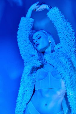 Çekici genç kadın seksi iç çamaşırları ve sahte kürkleriyle kolları başının üstünde ve gözleri kapalı poz veriyor.