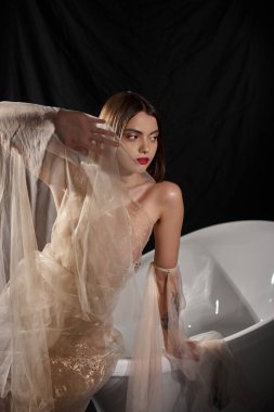 young woman with short hair posing in transparent dress posing near bathtub with black backdrop clipart