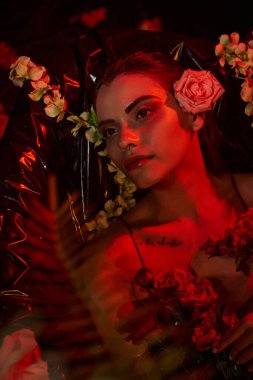 Farklı çiçeklerin arasında duran piercingli ve dövmeli çekici genç bir kadının yüzünde kırmızı ışık.