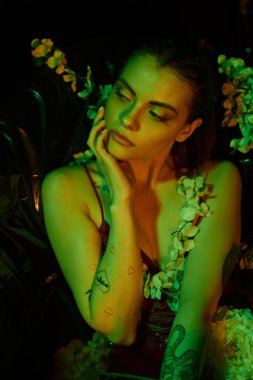 Yeşil ışık, ıslak saçlı çekici genç kadın yüzüne dokunuyor ve çiçeklerin arasında poz veriyor.