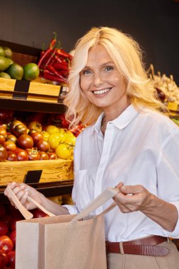 Normal giyinmiş olgun bir kadın. Arka planda market tezgahı olan açık bir alışveriş çantasıyla poz veriyor.