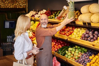 Neşeli sakallı satıcı marketin yanındaki bayan müşterisine kocaman ananas gösteriyor.