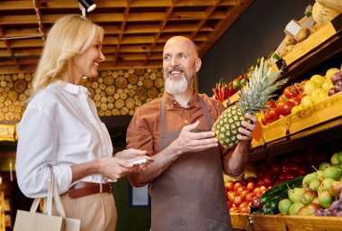 Sakallı, neşeli bayan müşterisine ananas gösteren olgun bir pazarlamacı.
