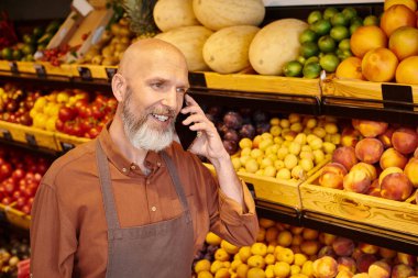 Olgun görünümlü, sakallı, markette çalışırken telefonla konuşan bir satıcı.