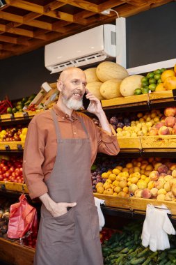 Gri sakallı olgun satıcı arka planda bakkal tezgahıyla el ele telefonla konuşuyor.