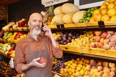 Neşeli gri sakallı satıcı telefonla konuşurken gülümsüyor ve elinde taze şeftali tutuyor.