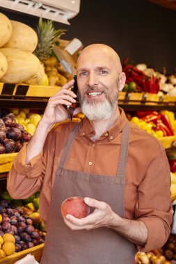 vertical shot of gray bearded jolly seller talking by phone and smiling at camera with peach in hand clipart