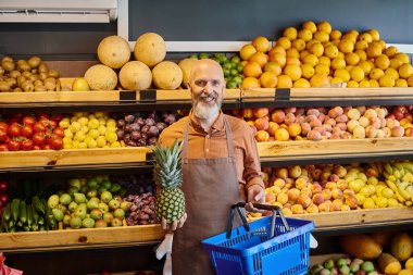 Neşeli iyi görünümlü satıcı elinde alışveriş sepeti, elinde ananas kameraya gülümsüyor.
