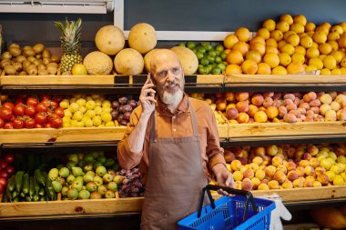 Gri sakallı olgun pazarlamacı elinde alışveriş sepetiyle markette telefonla konuşuyor.