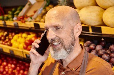 Neşeli olgun pazarlamacı arka planda bulanık market tezgahıyla telefonda aktif olarak konuşuyor.