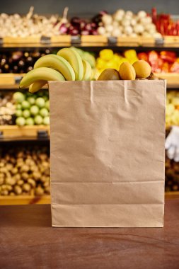 Arkaplanda market tezgahı olan, içi taze meyve dolu kocaman bir alışveriş çantasının nesnesi.