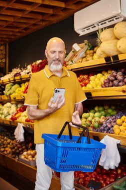 Neşeli sakallı müşteri alışveriş sepeti tutuyor ve markette telefonda neşeyle gülümsüyor.