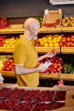 Gri sakallı, olgun bir müşteri elinde cep telefonuyla marketteki sulu domatese bakıyor.