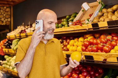 Neşeli gri sakallı adam marketten taze meyve seçerken cep telefonuyla konuşuyor.
