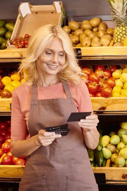 vertical shot of mature cheerful saleswoman smiling and looking at price tags at grocery store clipart
