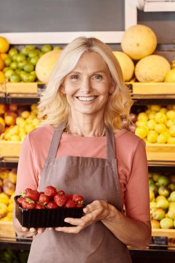 vertical shot of joyous mature seller holding pack of nutritious fresh strawberries smiling happily clipart