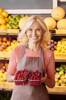 vertical shot of cheerful seller posing with packs of raspberries in hands and smiling at camera clipart