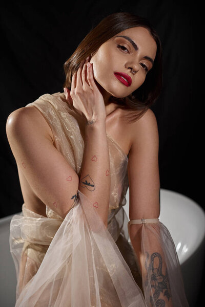 pretty woman with tattoos posing in transparent dress posing near bathtub with black backdrop