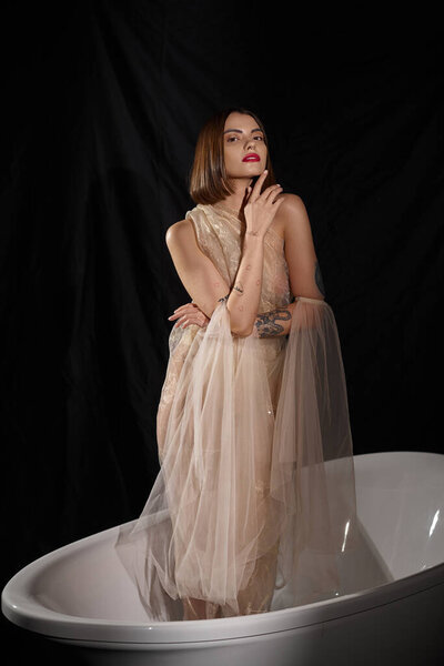 young woman with short hair posing in transparent dress posing in bathtub with black backdrop