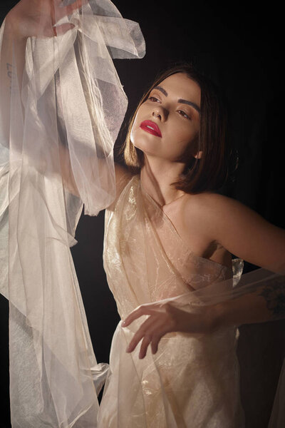 sensual young woman with short hair posing in transparent dress and gesturing on black backdrop