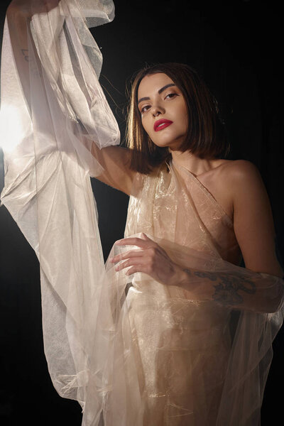 pretty young woman with short hair posing in transparent dress and gesturing on black backdrop