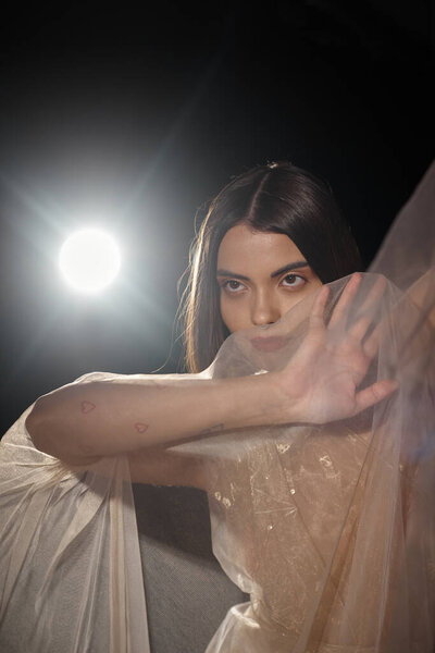 sensual young woman with short hair posing in transparent dress and gesturing on black backdrop