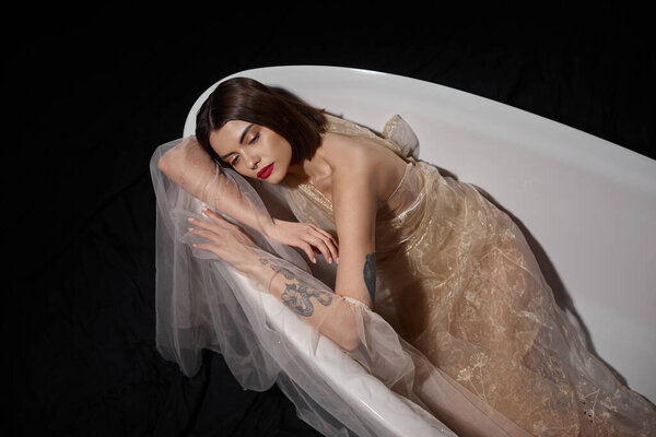 high angle view of graceful young woman in transparent dress while lying on bathtub on black