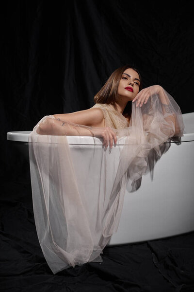 young graceful woman in transparent dress while sitting inside of bathtub on black backdrop