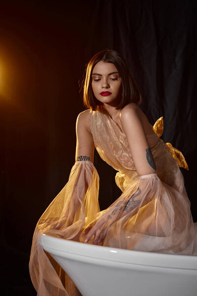 young graceful woman in romantic transparent dress standing near white bathtub on black backdrop