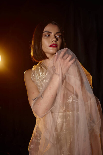 young graceful woman in transparent dress touching shoulder and looking away on black backdrop