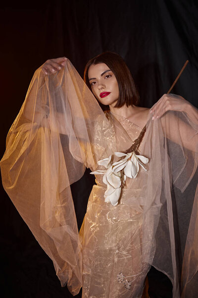 charming woman in romantic transparent dress holding white blooming flower on black backdrop