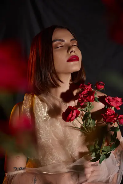 stock image dreamy young woman in romantic transparent dress holding blooming red roses on black backdrop