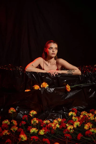 attractive young woman in wet slip dress sitting in black bathtub among beautiful flowers, red light
