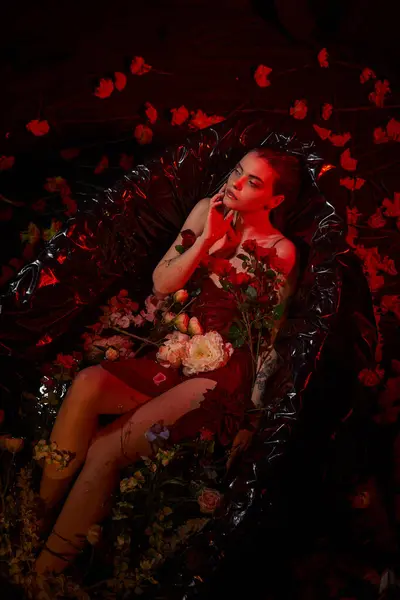 dreamy young woman in wet slip dress sitting in black bathtub with blooming flowers, red light