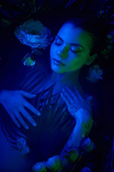 feminine beauty, beautiful young woman lying among palm leaves and flowers in water, blue light