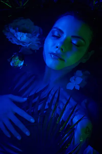 feminine beauty, relaxed young woman lying among palm leaves and flowers in water, blue light