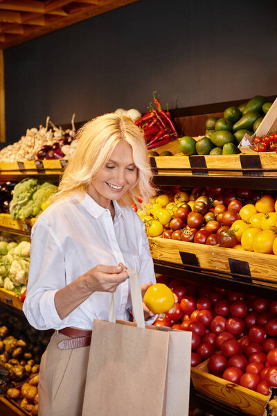 Весело зрелая женщина в повседневной одежде положить желтый помидор в сумку в то время как в продуктовом магазине
