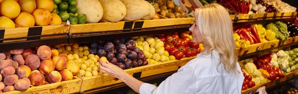 jolly mature woman in casual clothes with shopping bag choosing fruits at grocery store, banner