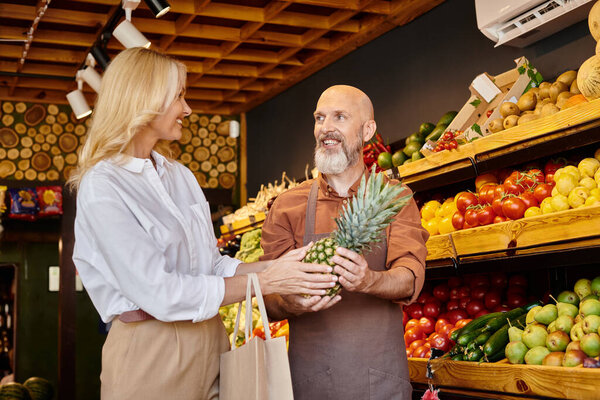 cheerful bearded seller giving fresh pineapple to his mature jolly female customer at grocery store