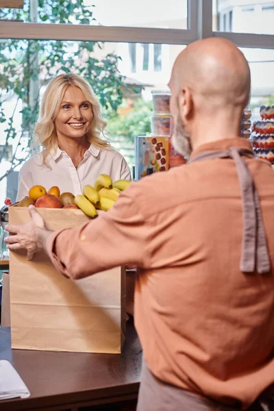 stock image focus on mature female customer smiling at blurred seller holding shopping bag with fruits