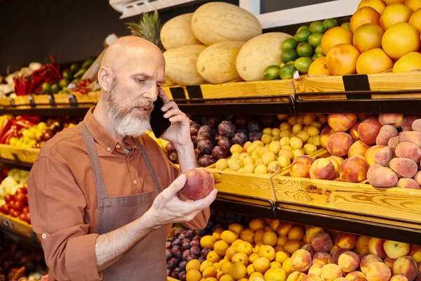 concentrated good looking bearded seller talking by mobile phone and looking at peach in his hand