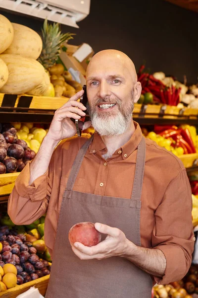 stock image vertical shot of gray bearded jolly seller talking by phone and smiling at camera with peach in hand