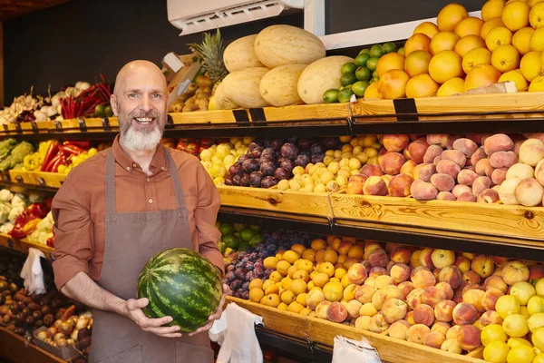 joyous good looking seller with beard posing with fresh watermelon in hands and smiling at camera