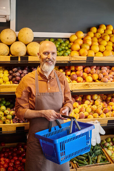 веселый зрелый продавец с корзиной в руках улыбаясь в камеру с фруктами на заднем плане