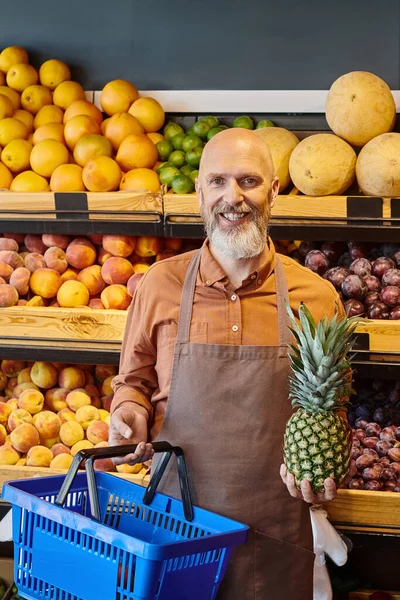 stock image vertical shot of jolly bearded salesman posing with shopping basket and fresh pineapple in hands