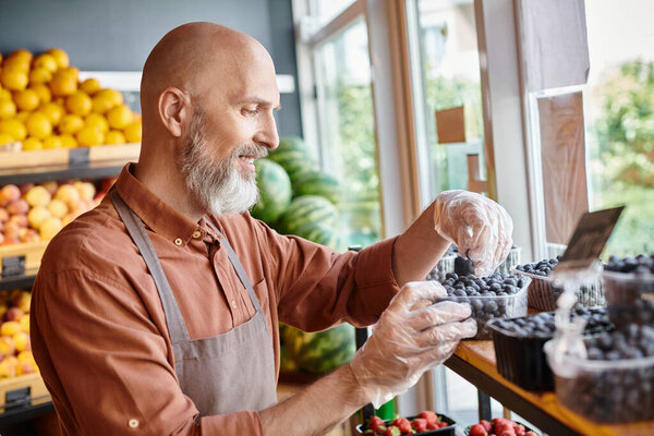 jolly bearded seller checking blueberries and smiling joyfully with fruits on stall on backdrop