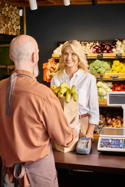 focus on blonde cheerful customer smiling at her blurred seller at cash desk with shopping bag on it