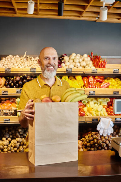 cheerful bearded man posing with paper bag full of fresh fruits and smiling at camera at grocery