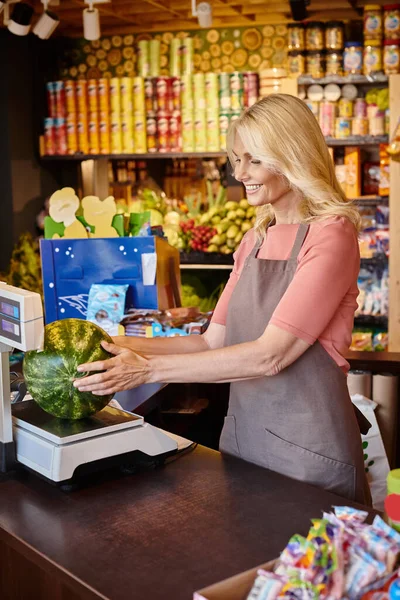 Neşeli olgun kadın satıcı markette taze karpuz tartıyor.